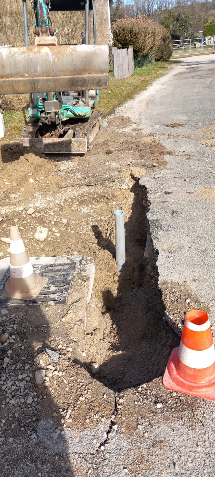 Tranchée étroite ouverte dans l'accotement en bordure de route, entre une mini-pelle et des cônes de signalisation. Un tuyau blanc émerge du fond de la fouille, près d'un regard bétonné.
