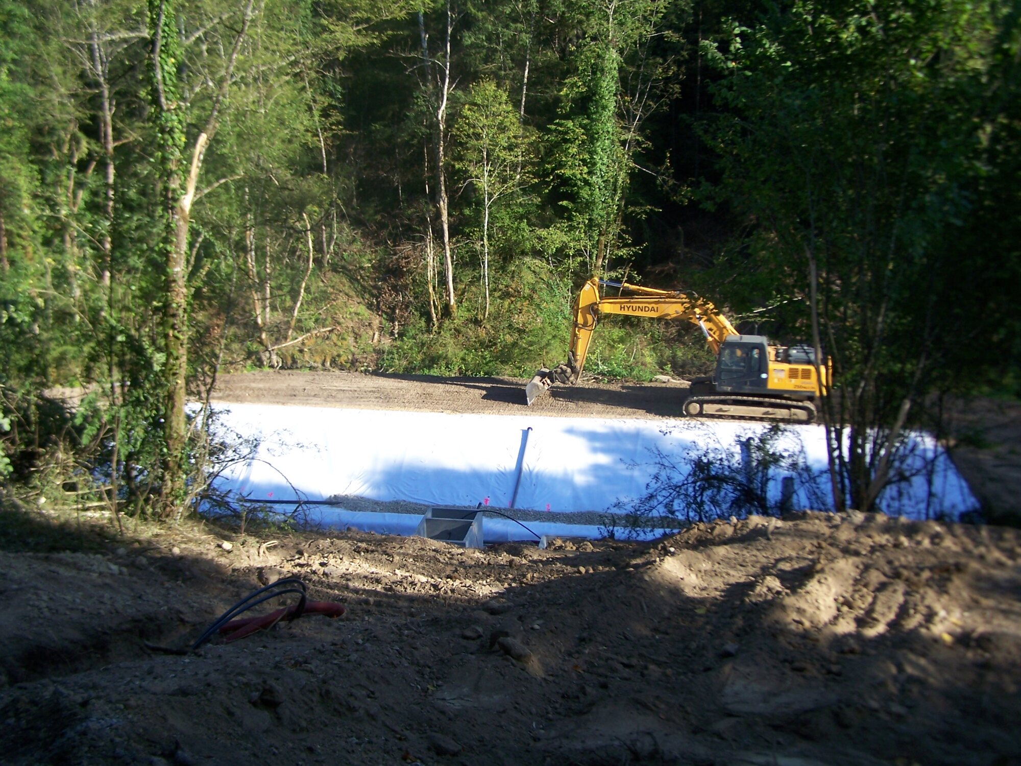 Chantier de la station de lagunage en lisière de forêt : bassin tapissé d'une bâche d'étanchéité claire, pelle mécanique Hyundai en cours de terrassement, terre remuée au premier plan.